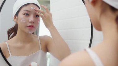 Genç ve güzel Asyalı bir kadın banyoda aynaya bakarken cildine krem sürüyor ya da losyon sürüyor. Tedavi için nemlendirici sürüyor. Yüz hatları, güzellik ve cilt bakımı için..