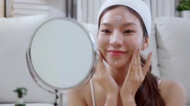 Güzel Asyalı bir kadın, oturma odasında aynaya bakarken kırışıklıklar için yüzüne krem sürüyor. Güzel bir kadın, yanağına nemlendirici sürüyor. Hijyen, cilt bakımı ve güzellik için..