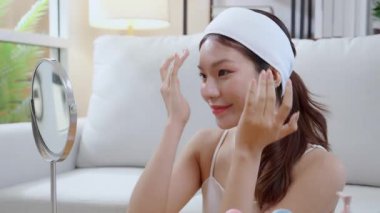 Güzel Asyalı bir kadın, oturma odasında aynaya bakarken kırışıklıklar için yüzüne krem sürüyor. Güzel bir kadın, yanağına nemlendirici sürüyor. Hijyen, cilt bakımı ve güzellik için..