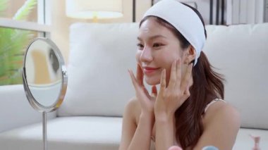Güzel Asyalı bir kadın, oturma odasında aynaya bakarken kırışıklıklar için yüzüne krem sürüyor. Güzel bir kadın, yanağına nemlendirici sürüyor. Hijyen, cilt bakımı ve güzellik için..