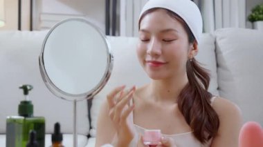 Güzel Asyalı bir kadın, oturma odasında aynaya bakarken kırışıklıklar için yüzüne krem sürüyor. Güzel bir kadın, yanağına nemlendirici sürüyor. Hijyen, cilt bakımı ve güzellik için..