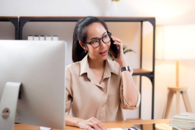 Genç Asyalı iş kadını ofis telefonuyla konuşurken masaüstü bilgisayarı kullanıyor, iş kadını telefon, iş ya da danışmanlık yaparken masaüstü bilgisayarıyla çalışıyor.