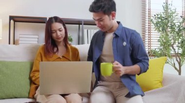 Genç Asyalı çift rahat ve rahat bir şekilde dizüstü bilgisayara bakıyor. Oturma odasındaki kanepede oturuyor. Kanepede dizüstü bilgisayar izliyor..
