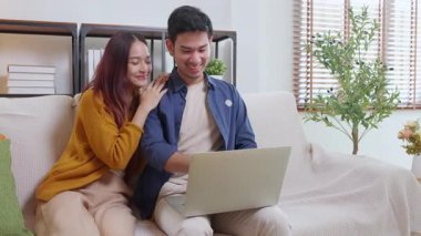 Genç Asyalı çift rahat ve rahat bir şekilde dizüstü bilgisayara bakıyor. Oturma odasındaki kanepede oturuyor. Kanepede dizüstü bilgisayar izliyor..