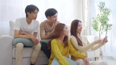 Genç Asyalı arkadaşlar evdeki oturma odasında dijital tablet kullanarak selfie ya da video çekiyorlar. Sosyal medya, etkileşim ve arkadaşlıkla bağlantı kurarken neşeli ve gülümsüyorlar..