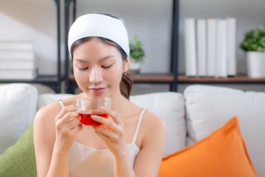 Saç bandı takmış, çay içen güzel Asyalı bir kadın. Oturma odasındaki koltukta oturmuş çay içiyor. Mutlu bir kadın, sağlık ve rahatlama için çay içiyor..