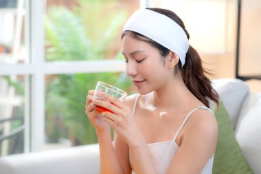 Saç bandı takmış, çay içen güzel Asyalı bir kadın. Oturma odasındaki koltukta oturmuş çay içiyor. Mutlu bir kadın, sağlık ve rahatlama için çay içiyor..