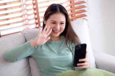 Genç Asyalı bir kadın kanepede oturmuş videolu telefon bekliyor. Oturma odasında telefon görüşmesi yapıyor. Sosyal medyayı internet üzerinden sohbet etmek için kullanıyor..