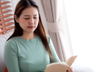 Genç Asyalı bir kadın, yüz ifadesiyle kitap okuyor. Oturma odasındaki koltukta oturuyor, kadın rahatlıyor ve kanepeye oturuyor. Edebiyat, etkinlik ve hobi, yaşam tarzı ve rahatlama okuyor..