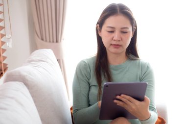Genç Asyalı iş kadını rahatlamak için tablet bilgisayar kullanıyor ve sosyal medyayı kontrol ediyor. Ev ofisinin oturma odasında kanepede oturuyor. İş kadını tabletle çalışıyor..