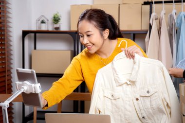 İki genç Asyalı kadın online giyim mağazasında yaşarken, evde sosyal medyada tablet ve dizüstü bilgisayarlı giysiler gösteriliyor. Girişimci çevrimiçi kıyafet satışı ve sunumu için çevrimiçi satış yapıyor..