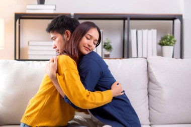 Genç Asyalı çift, evdeki oturma odasında sevgi dolu bir şekilde kanepede otururken birbirlerine sarılırlar. Karı koca, nazik ve sevgi dolu bir şekilde birbirlerine sarılırlar. Birbirlerine sarılırlar..