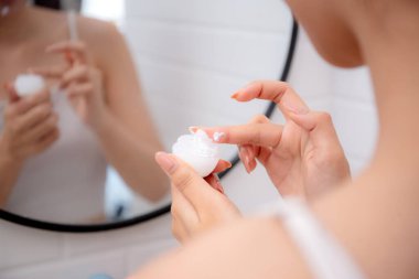 Genç bir kadının banyoda aynaya bakarken cildine krem sürmesi ya da losyon sürmesi, tedavi için nemlendirici sürmesi, yüz, güzellik ve cilt kırışıklıkları..