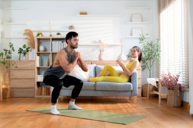 Genç Asyalı bir adam evdeki yoga minderinde yoga yaparken kadınlar koltukta dinlenmek, spor yapmak ve sağlıklı bir yaşam tarzını rahat, güçlü ve güçlü bir kas gücüyle kullanırlar..
