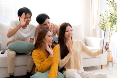 Genç Asyalı arkadaşlar evdeki oturma odasında dijital tablet kullanarak selfie ya da video çekiyorlar. Sosyal medya, etkileşim ve arkadaşlıkla bağlantı kurarken neşeli ve gülümsüyorlar..