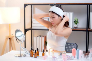 Saç bandı takan ve saç bağlayan Asyalı genç bir kadın evde aynaya bakarken, güzellik ve güzellik masasında kozmetik ürünler, güzel bir kadın ve kadınlık..