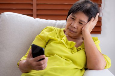 Kıdemli Asyalı kadın evdeki akıllı telefona bakarken koltukta oturup endişeleniyor ve strese giriyor. Mutsuz ve endişeli bir kadın akıllı telefondan, yaşam tarzından ve iletişimden izliyor..