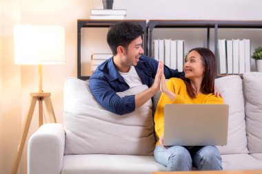 Mutlu Asyalı çift evdeki oturma odasında kanepede oturmuş laptopla çalışırken başarıyı kutluyor, gülümsüyor ve takım çalışmasından zevk alıyor, uzaktan işbirliği yapıyor, birlikte neşe içinde..