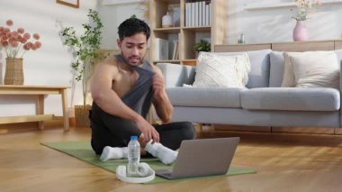 Evde antreman yaptıktan sonra yoga minderinde oturan yorgun Asyalı genç adam oturma odasında su içiyor. Egzersiz yaptıktan sonra rahat, özgüvenli ve rahat bir şekilde dizüstü bilgisayar izliyor..