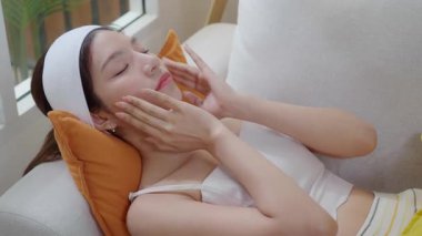 Genç Asyalı kadın kanepede uzanmış yüz masajı yaparken cilt bakımlı güzellik, rahatlık ve huzur içinde rahat, güzel bir kadın ve şımarık bir refah içinde..