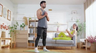 Genç Asyalı bir adam evdeki yoga minderinde yoga yaparken kadınlar koltukta dinlenmek, spor yapmak ve sağlıklı bir yaşam tarzını rahat, güçlü ve güçlü bir kas gücüyle kullanırlar..