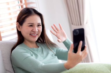 Genç Asyalı bir kadın kanepede oturmuş videolu telefon bekliyor. Oturma odasında telefon görüşmesi yapıyor. Sosyal medyayı internet üzerinden sohbet etmek için kullanıyor..