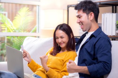 Mutlu Asyalı çift evdeki oturma odasında birlikte online alışveriş yapmak için dizüstü bilgisayar ve kredi kartı kullanıyor, kadın ve erkek gülümsüyor ve dijital yaşam tarzının keyfini çıkarıyor, uygun bir ödeme.