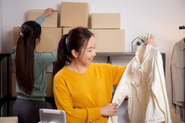 İki genç Asyalı kadın online giyim mağazasında yaşarken, evde sosyal tabletle giysi satışı yaparken, girişimci çevrimiçi kıyafet satışı ve sunumu için çevrimiçi satış yapıyor..