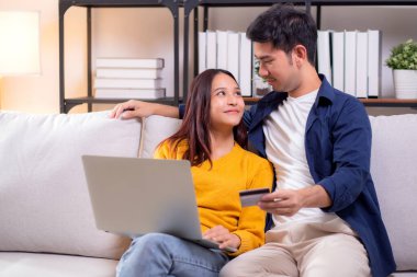 Mutlu Asyalı çift evdeki oturma odasında birlikte online alışveriş yapmak için dizüstü bilgisayar ve kredi kartı kullanıyor, kadın ve erkek gülümsüyor ve dijital yaşam tarzının keyfini çıkarıyor, uygun bir ödeme.