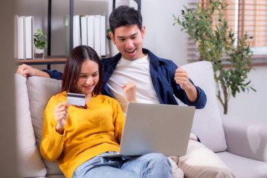 Mutlu Asyalı çift evdeki oturma odasında birlikte online alışveriş yapmak için dizüstü bilgisayar ve kredi kartı kullanıyor, kadın ve erkek gülümsüyor ve dijital yaşam tarzının keyfini çıkarıyor, uygun bir ödeme.
