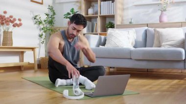 Evde antreman yaptıktan sonra yoga minderinde oturan yorgun Asyalı genç adam oturma odasında su içiyor. Egzersiz yaptıktan sonra rahat, özgüvenli ve rahat bir şekilde dizüstü bilgisayar izliyor..