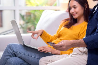 Yakın plan mutlu Asyalı çift evdeki online alışveriş için dizüstü bilgisayar ve kredi kartı kullanıyor. Kadın ve erkek, dijital bir yaşam tarzı ve uygun bir ödeme ile rahatlıyor..