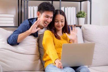Mutlu Asyalı çiftler, dizüstü bilgisayarlarında oturma odasındaki koltukta oturup rahatlama, dizüstü bilgisayar, yaşam tarzı ve etkileşimi izleyen çiftlerle çevrimiçi iletişim kurarken gülümsüyor ve el sallıyorlar..