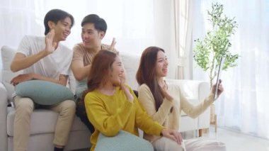 Genç Asyalı arkadaşlar evdeki oturma odasında dijital tablet kullanarak selfie ya da video çekiyorlar. Sosyal medya, etkileşim ve arkadaşlıkla bağlantı kurarken neşeli ve gülümsüyorlar..