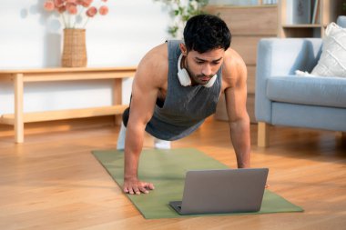 Genç Asyalı adam evde antrenman yapıyor. Evde dizüstü bilgisayarla ev bilgisayarı izliyor. Oturma odasında, sağlık, sağlıklı yaşam tarzı ve egzersiz için vücut gücü saptadı ve motive etti..
