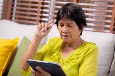 Kıdemli Asyalı kadın kanepeye oturmuş dijital tablet kullanıyor. Oturma odasında dinleniyor, yaşlılar tablete oturmuş düşünceli, rahat bir hayat tarzı ve iletişim kuruyor..