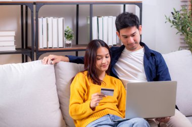 Mutlu Asyalı çift evdeki oturma odasında birlikte online alışveriş yapmak için dizüstü bilgisayar ve kredi kartı kullanıyor, kadın ve erkek gülümsüyor ve dijital yaşam tarzının keyfini çıkarıyor, uygun bir ödeme.