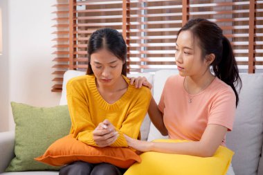 Genç Asyalı bir kadın, arkadaşını ağlama ve depresyonla rahatlatırken oturma odasında kanepede oturuyor, arkadaşlığın sıkıntısı ve empatisi, umutsuzluk ve ruh sağlığını desteklerken, bir kadın da arkadaşını teşvik ediyor..