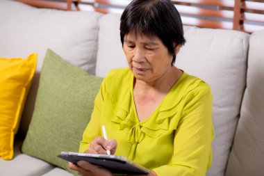 Kıdemli Asyalı kadın kanepeye oturmuş dijital tablet kullanıyor. Oturma odasında dinleniyor, yaşlılar koltukta oturuyor. Tablete yazılar yazıyor..