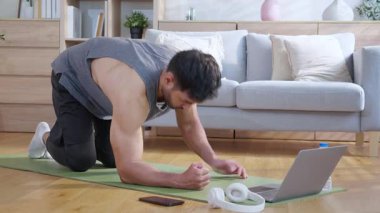 Genç Asyalı adam evde oturma odasında kalas egzersizi yaparken dizüstü bilgisayarda dayanıklılık ve motivasyonla antrenman eğitimi alırken yoga minderinde adam eğitim tahtası, dijital fitness yaşam tarzı..