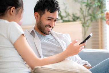 Genç Asyalı çift evdeki oturma odasında birlikte otururken rahat bir yaşam tarzı ve bağ kurma, ilişki ve boş vakit geçirme, dijital içeriği mutlu bir şekilde paylaşma ve keyif alma..