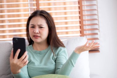 Genç Asyalı kadın evdeki akıllı telefona bakarken kanepede oturmuş endişeli ve stresli bir şekilde, mutlu olmayan ve endişeli bir kadın akıllı telefondan, yaşam tarzından ve iletişimden izliyor..