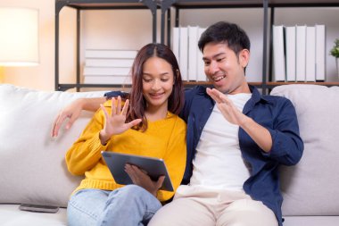 Mutlu Asyalı çift videosu tablet üzerinde oturma odasındaki koltukta oturuyor, gülümsüyor ve internette rahatlama, tablet, yaşam tarzı ve etkileşim izlerken el sallıyor..