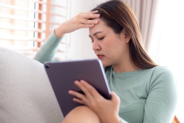 Tablet bilgisayar kullanan genç Asyalı iş kadını ev ofisinin oturma odasındaki kanepede otururken endişeli, iş kadını endişeli, yorgun, iş ve yaşam tarzını düşünürken tablet üzerinde çalışıyor..
