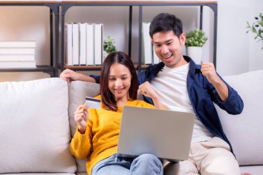 Mutlu Asyalı çift evdeki oturma odasında birlikte online alışveriş yapmak için dizüstü bilgisayar ve kredi kartı kullanıyor, kadın ve erkek gülümsüyor ve dijital yaşam tarzının keyfini çıkarıyor, uygun bir ödeme.