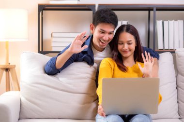 Mutlu Asyalı çiftler, dizüstü bilgisayarlarında oturma odasındaki koltukta oturup rahatlama, dizüstü bilgisayar, yaşam tarzı ve etkileşimi izleyen çiftlerle çevrimiçi iletişim kurarken gülümsüyor ve el sallıyorlar..