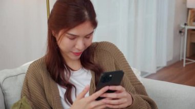 Modern evdeki oturma odasında telefonla konuşurken gülümseyen Asyalı genç kadın, yaşam tarzını rahatlat, konuşmaktan zevk alan mutlu kadın, neşeli yüz ifadesi, eğlenceli telefon konuşması yapan mutlu kadın..