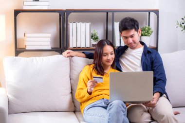 Mutlu Asyalı çift evdeki oturma odasında birlikte online alışveriş yapmak için dizüstü bilgisayar ve kredi kartı kullanıyor, kadın ve erkek gülümsüyor ve dijital yaşam tarzının keyfini çıkarıyor, uygun bir ödeme.
