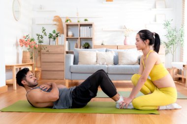 Genç Asyalı çift evde oturma odasında antrenman yaparken, kadın da yoga minderindeki abdominal mekik sırasında motivasyon, sağlık ve hedefler için fitness ve takım çalışması ile yardımcı oluyor..