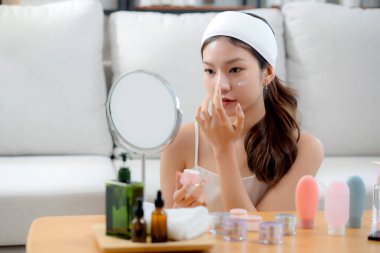 Güzel Asyalı bir kadın, oturma odasında aynaya bakarken kırışıklıklar için yüzüne krem sürüyor. Güzel bir kadın, yanağına nemlendirici sürüyor. Hijyen, cilt bakımı ve güzellik için..